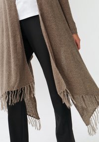Cárdigan largo y abierto en suave tejido de punto color marrón. Presenta flecos en el bajo y mangas largas, combinado con pantalones negros.