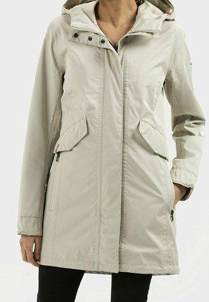 Vaaleanbeige huppari sadetakki, jossa edessä vetoketju, napillinen tuulisuojaläppä, läppätaskut ja sivussa vetoketjutaskut, päällä henkilö, jonka toisessa kädessä on taskussa.