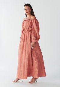 Robe maxi en corail, design décolleté avec taille élastique, manches longues bouffantes et tissu léger texturé. Stylisée avec des talons blancs.