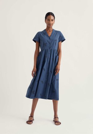 Žena oblečená v modrých midi šatách na gombíky s krátkym rukávom a kvetinovým vzorom, stojaca v hnedých sandáloch na jednofarebnom pozadí.