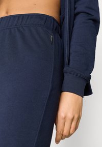 Αθλητική ένδυση σε ναυτικό μπλε με ελαστική μέση και μανίκια με μανσέτες, κατασκευασμένη από υφασμάτινο με υφή και διακριτική λεπτομέρεια λογότυπου.