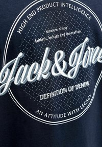 Marineblauw T-shirt met witte tekst, cirkelvormig logo en diamantenpatroon. Glad materiaal met een casual ontwerp.