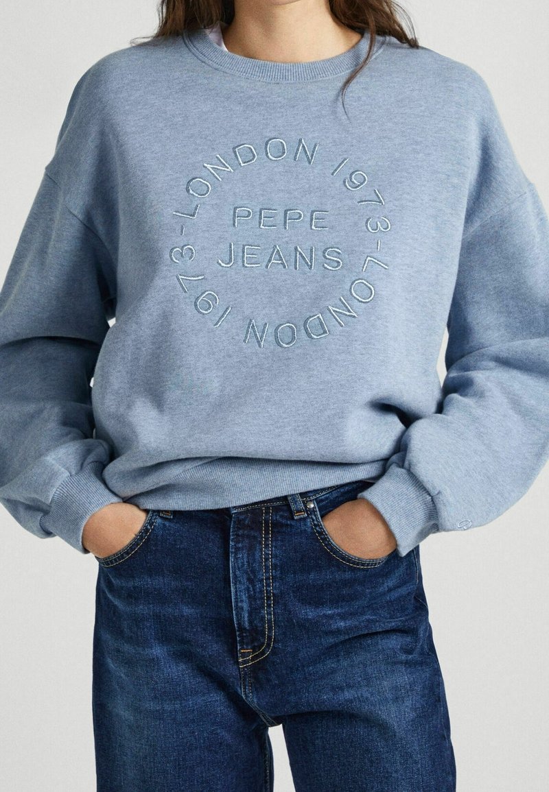 Personne portant un sweat-shirt bleu avec « Pepe Jeans » et « London 1973 » brodés en cercle, associé à un jean bleu foncé. Mains dans les poches.