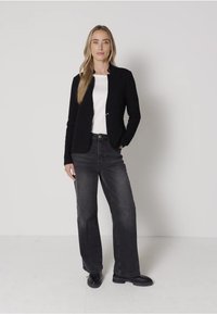 Musta räätälöity bleiseri valkoisen paidan päällä, yhdistettynä korkeavyötäröisiin harmaisiin farkkuihin ja mustiin saappaisiin. Silkkinen kangas ja tiukka muotoilu.