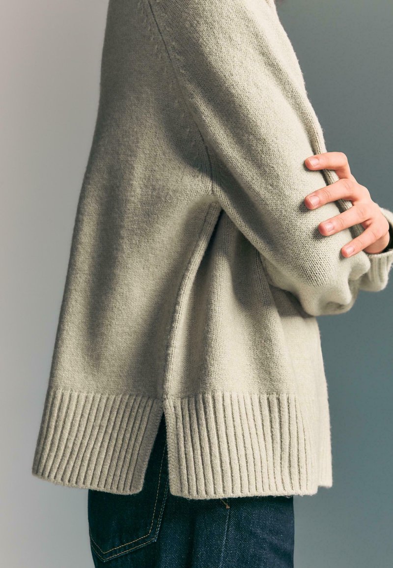 Persona con le braccia incrociate che indossa un maglione lavorato a maglia beige chiaro con orlo a costine e jeans blu scuro, su uno sfondo neutro.