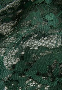 Tissu en dentelle vert foncé avec des motifs floraux et feuillus, montrant une texture détaillée des fils et des zones de maille ouvertes.