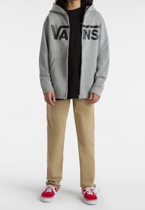 Sweat à capuche gris zippé avec le logo "VANS" noir, associé à un t-shirt noir, un pantalon beige et des baskets rouges, offrant une coupe décontractée et une capuche.