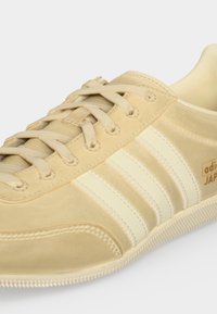 Μπεζ αθλητικό παπούτσι Adidas Japan με άσπρες ρίγες, μπεζ κορδόνια και υφή στη σόλα, εμφανίζεται από κοντινή γωνιακή λήψη.