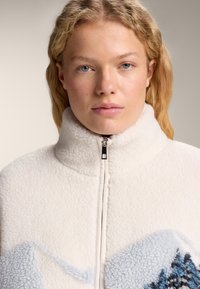 Femeie blondă cu ochi albaștri, purtând o jachetă albă din fleece cu guler înalt și model albastru de munți, pe un fundal neutru.