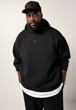 Uomo con barba che indossa un hoodie nero e un cappellino da baseball nero con pantaloni bianchi, in piedi con le mani nelle tasche su uno sfondo neutro.