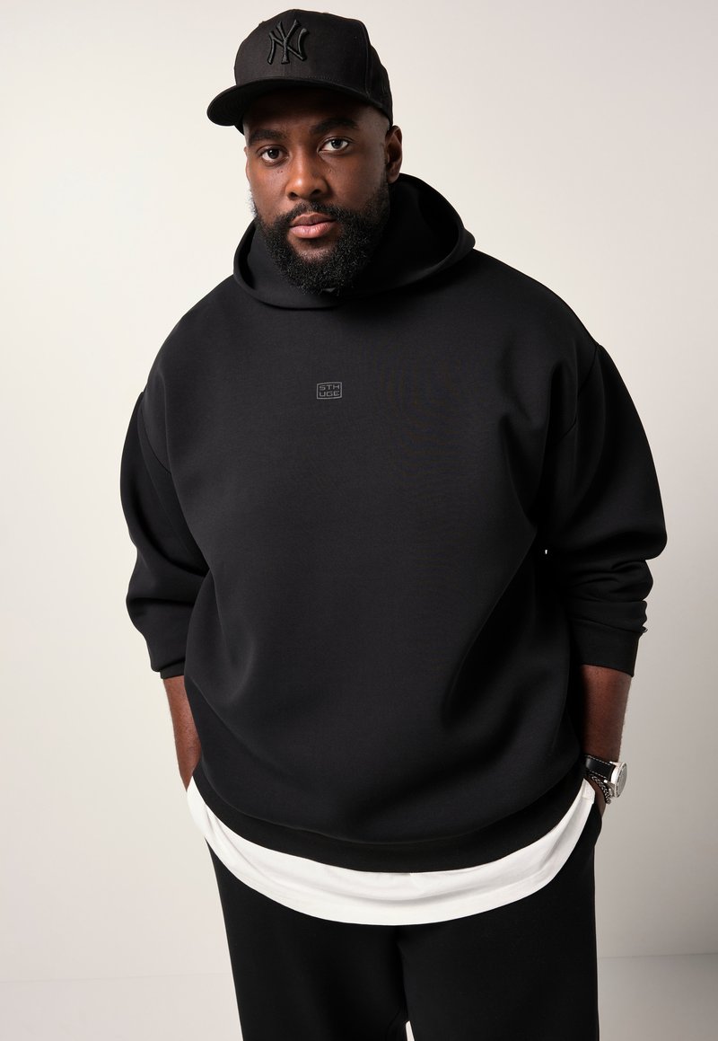 Uomo con barba che indossa un hoodie nero e un cappellino da baseball nero con pantaloni bianchi, in piedi con le mani nelle tasche su uno sfondo neutro.