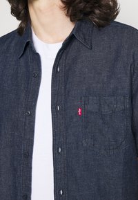 Mörk denimskjorta med knappar och en subtil textur, med en bröstficka och en liten röd logotagg på vänster sida.