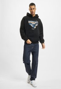 Felpa nera con graphic colorato di un'aquila e testo sul davanti, abbinata a jeans blu scuro e sneakers bianche. Tessuto morbido, vestibilità casual.