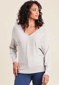 Helles graues, glitzerndes Sweater mit V-Ausschnitt, überschnittenen Schultern und Knopfdetails an den Ärmeln, kombiniert mit dunklen Jeans. Seidig glatte Textur.