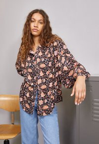 Blusa negra con flores naranjas y rosas, de corte holgado, con botones en la parte delantera y mangas amplias. Combinada con jeans de mezclilla claros.
