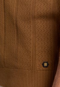 Bruine gebreide trui met een textuurpatroon, met verticale strepen en perforaties. Bevat een zwarte hexagonale logo-label aan de onderkant.