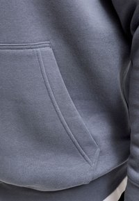 Gros plan d'une personne portant un sweat-shirt gris avec une poche kangourou à l'avant et des détails de couture visibles.