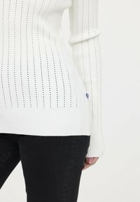 Weißer gerippter Strickpullover mit horizontalen und vertikalen Streifenmustern, langen Ärmeln, figurnaher Silhouette und einem kleinen Logoetikett am Saum.