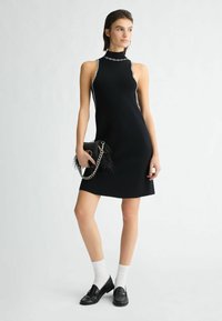 Abito nero senza maniche con scollatura alta, dettagli in catena argentata lungo i lati, abbinato a una clutch nera e mocassini neri.