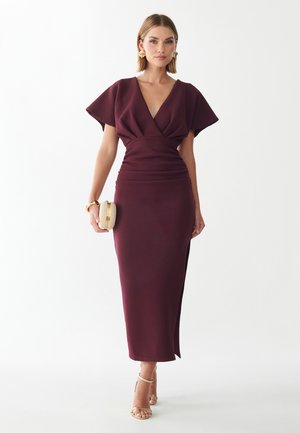 Donna che indossa un abito midi bordeaux con profondo scollo a V, maniche larghe, orecchini dorati, con una pochette beige e sandali con cinturini.