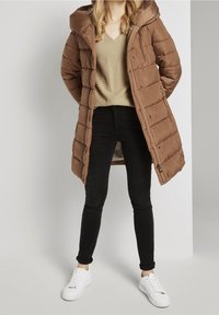 Brun quiltet pufferjakke med hætte, med trykknapper, båret over en struktureret beige top og sorte jeans, parret med hvide sneakers.