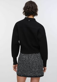 Pullover nero con collo e maniche oversize, abbinato a una gonna con motivo a quadretti bianco e nero con dettagli a frange lungo l'orlo.