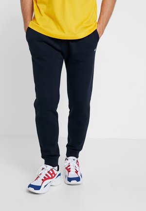 Muž stojící s rukama v kapsách, oblečený v černých teplákových kalhotách, žlutém tričku a bílých teniskách s červenými a modrými akcenty.