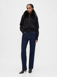 Pull noir en maille à fermeture éclair avec un col en fausse fourrure, associé à un jean en denim bleu à jambes droites et des bottines noires à talons.