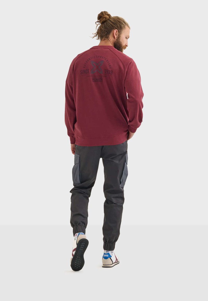 Felpa rossa a maniche lunghe con testo stampato sul retro, abbinata a pantaloni cargo grigi scuri con tasche laterali e polsi elasticizzati.