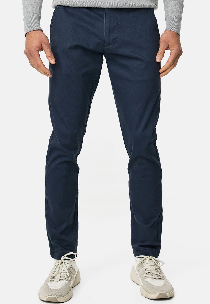 Mann trägt marineblaue Slim-Fit-Hosen, grauen Pullover und hellgraue Sportschuhe, steht vor weißem Hintergrund.