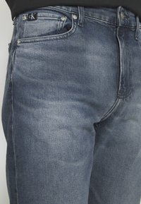 Jeans de denim azul oscuro con un lavado deslavado, diseño de cinco bolsillos y un logo de CK en la cinturilla. Presenta herrajes plateados y acentos cosidos.