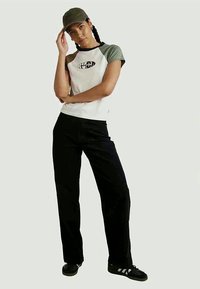 Bílý tričko s krátkými zelenými rukávy a grafickým logem, spárované s černými kalhotami a černými teniskami. Zelená čepice dotváří celý outfit.