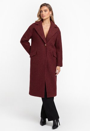 Donna che indossa un lungo cappotto di lana rosso scuro sopra pantaloni neri e stivali neri con tacco, in piedi con una mano sul fianco su sfondo bianco.