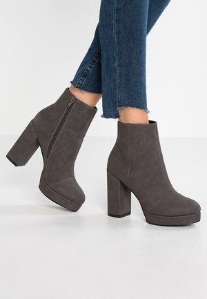 Bottines à talons hauts grises avec une finition lisse, fermeture éclair latérale, semelle épaisse et un talon carré, associées à un jean à ourlet effiloché.