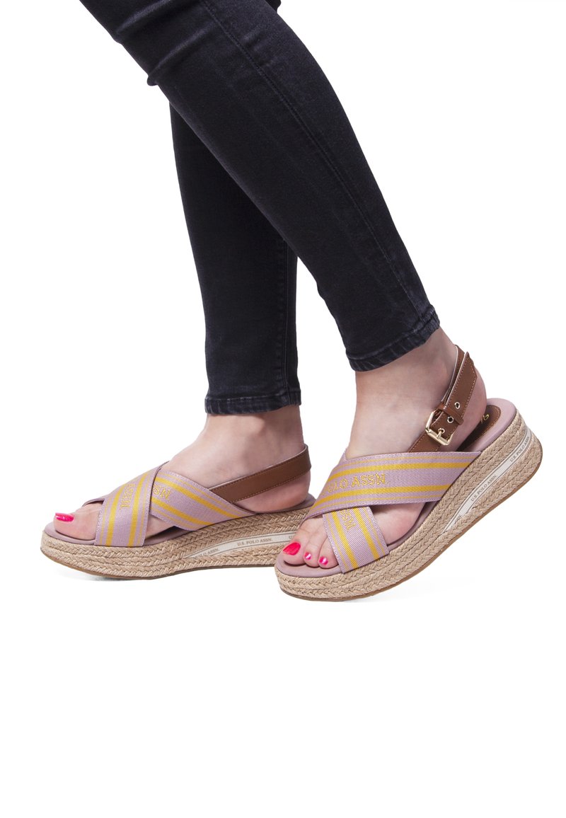 Noge v wedges sandalu s svetlo rjavimi in vijoličnimi križnimi trakovi, z zlatim zaponko in rožnato manikuro nohtov, v kombinaciji s črnimi oprijetimi jeans.