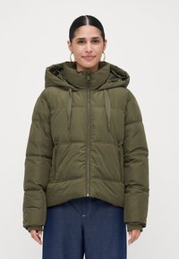 Vrouw in een olijfgroene gewatteerde capuchonjas, dichtgeritst, met ontspannen handen langs de zijkanten en donkerblauwe jeans tegen een effen achtergrond.