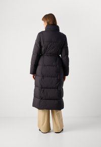 Άτομο που φοράει ένα μακρύ μαύρο παλτό με επένδυση και ψηλό γιακά, με τα χέρια χαλαρά δίπλα στα πλάγια, μπεζ παντελόνι, στέκεται μπροστά σε εντελώς λευκό φόντο.