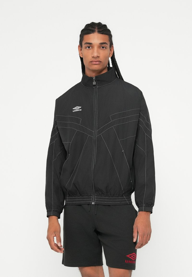 Mladic s dugim pletenicama nosi crnu zip-up jaknu Umbro i crne Umbro kratke hlače na bijeloj pozadini.
