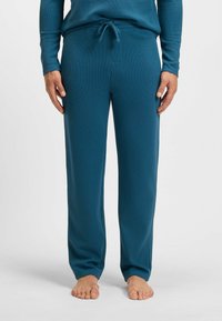 Homme portant un pantalon de détente texturé teal avec taille à cordon, debout pieds nus sur un fond blanc uni.