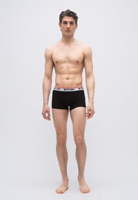 Νεαρός άνδρας όρθιος, ξυπόλυτος, φορώντας μαύρο μπόξερ Moschino, μπροστά σε απλό λευκό φόντο.