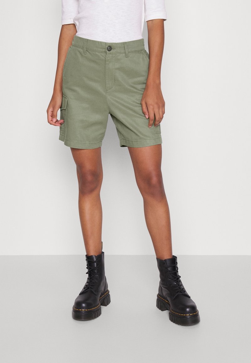 Pantaloni cargo in cotone verde oliva con chiusura a bottone, due tasche laterali e orlo arrotolato, abbinati a stivaletti da combattimento neri con lacci.