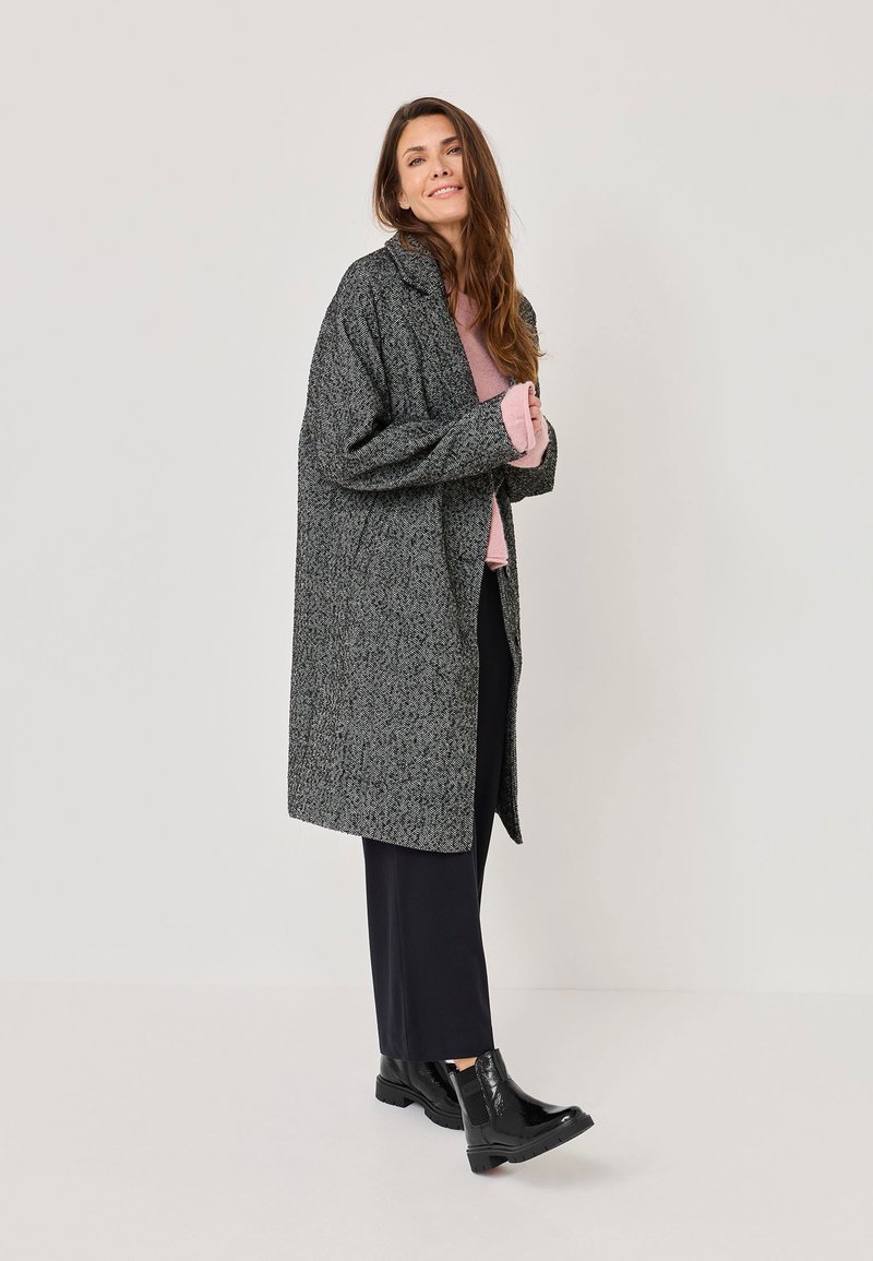Cappotto grigio oversize con un motivo testurizzato, frontale aperto e colletto alla cinese, abbinato a un maglione rosa e stivaletti neri alla caviglia.