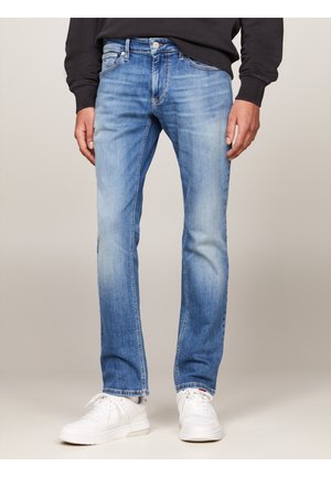 Mann trägt hellblaue ausgewaschene gerade Jeans, weiße Sneaker und ein schwarzes langärmeliges Shirt, steht vor neutralem Hintergrund.