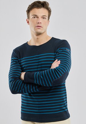 Jeune homme à la peau claire et aux cheveux bruns portant une chemise à manches longues rayée bleu marine et sarcelle, debout les bras croisés devant un fond uni.