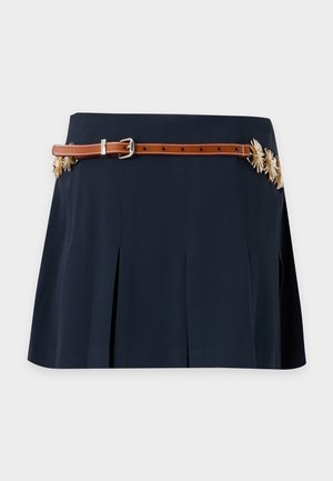 Femme portant une veste courte à manches courtes bleu marine avec bordure florale, une blouse blanche, une mini-jupe plissée bleu marine, une ceinture marron et des talons hauts beiges.