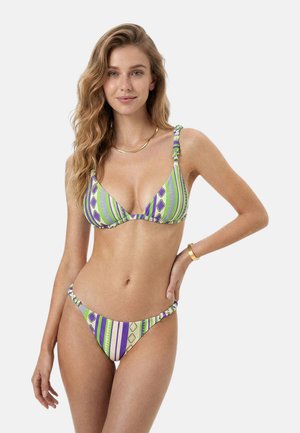 Giovane donna con capelli biondi ondulati che indossa un bikini a righe multicolore con motivi geometrici, collana d'oro e bracciale, posando con la mano sul fianco.