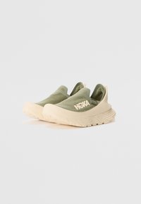 Green slip-on sneakers with a textured upper, beige sole, and HOKA branding on the side. Lightweight design with a sporty silhouette.