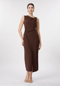 Γυναίκα που φοράει αμάνικο, καφέ σκούρο midi φόρεμα με λεπτομέρειες με αγκράφες στο πλάι και μπεζ σανδάλια με λουράκια, σε λευκό φόντο.