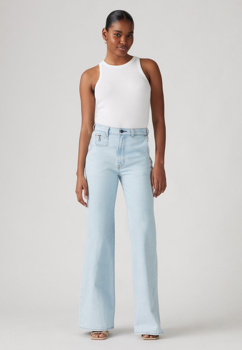 Jeans évasés bleu clair à taille haute, fabriqués en denim. Associés à un top en débardeur blanc ajusté. Texture lisse, détails minimalistes.