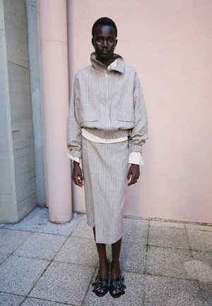Black person stands against pink wall, wearing beige checkered zip jacket and matching midi skirt, with black open-toe buckle shoes outdoors.